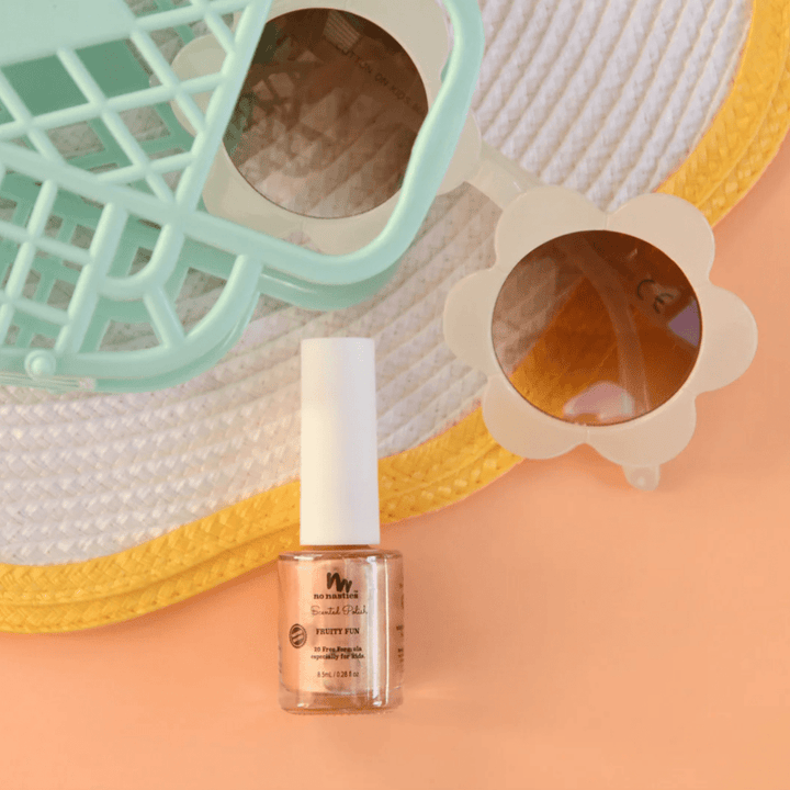 A bottle of No Nasties 20 FREE Kids Scented Nail Polish rests on a peach surface, next to a pair of sunglasses with flower-shaped frames and a green plastic basket against the backdrop of a white and yellow hat.