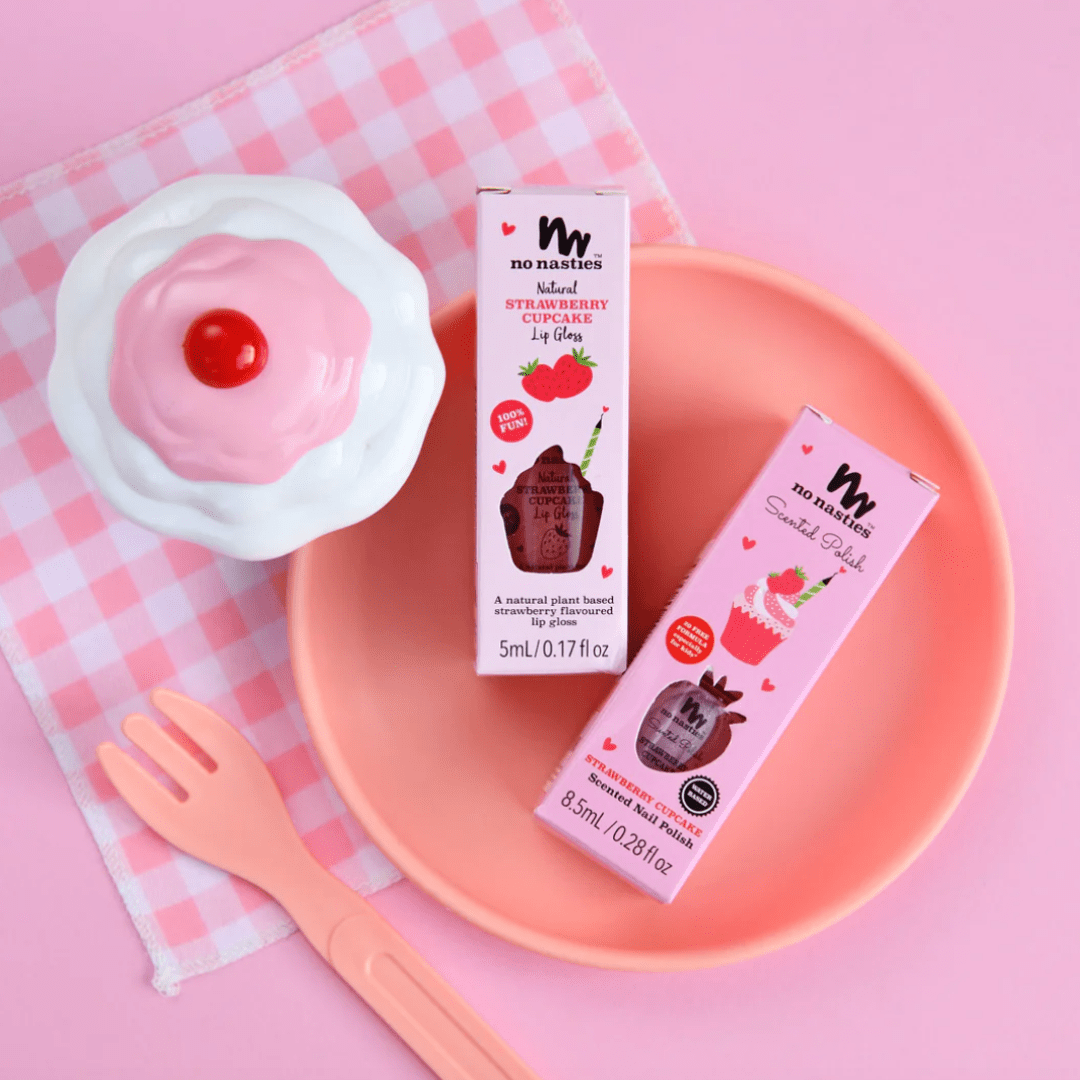 Two boxes of No Nasties lip gloss placed in a pink bowl, accompanied by a toy cupcake, No Nasties 20 FREE Kids Scented Nail Polish, and a pink fork on a pink checkered cloth.