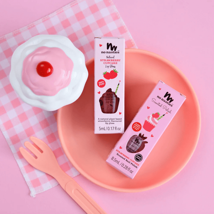 Two boxes of No Nasties lip gloss placed in a pink bowl, accompanied by a toy cupcake, No Nasties 20 FREE Kids Scented Nail Polish, and a pink fork on a pink checkered cloth.