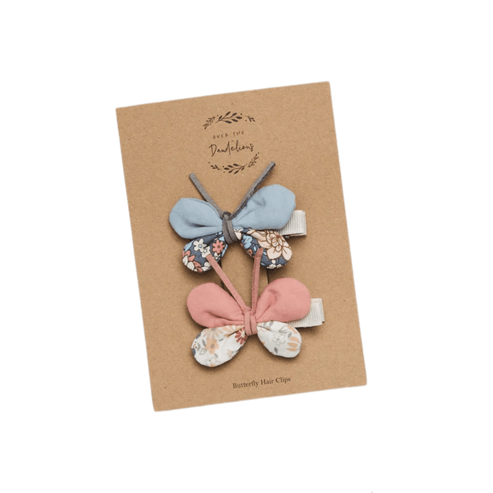 Two butterfly-shaped hair clips, one blue and one pink, attached to a brown cardboard backing with text that reads "Over the Dandelions." Perfect for toddlers, these Over the Dandelions Butterfly Hair Clips by Over the Dandelions add a charming touch to any outfit.