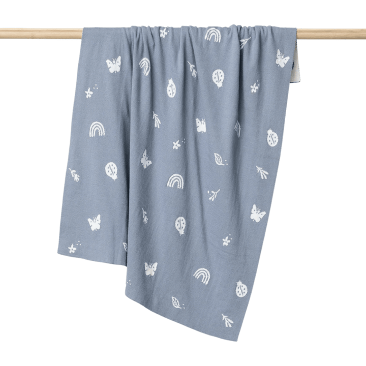 The Over the Dandelions Organic Cotton Print Blanket (Multiple Variants) in light blue showcases a delightful white pattern of butterflies, rainbows, leaves, and other small designs. This charming knitted baby blanket is beautifully displayed hanging on a wooden rod.