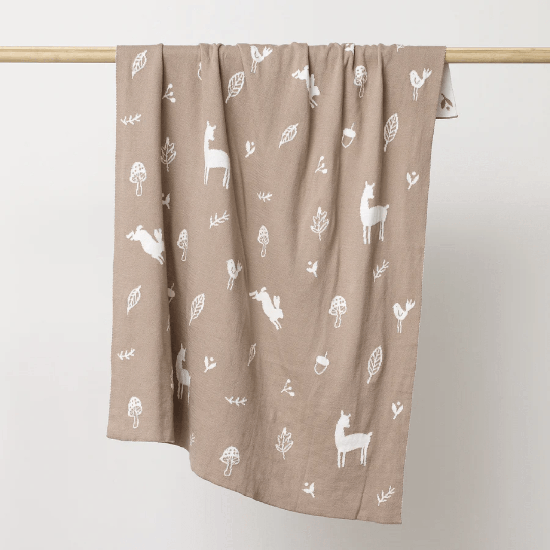 This delightful nursery decor item, the Over the Dandelions Organic Cotton Print Blanket, features white animal and nature patterns on a beige fabric. It hangs gracefully on a wooden rod, resembling an elegant addition to your baby's room.