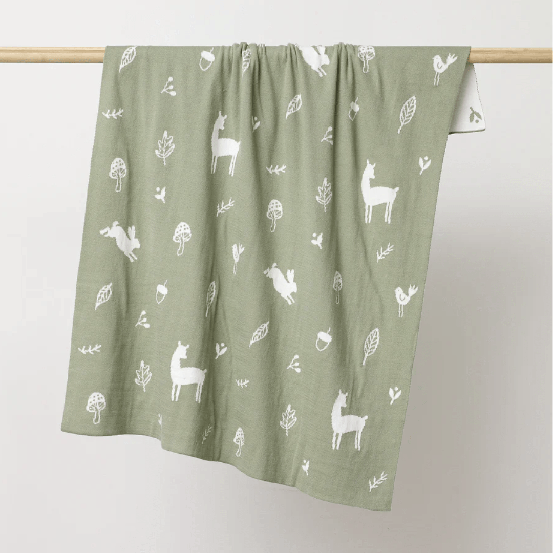 Hanging on a wooden rod is an Over the Dandelions Organic Cotton Print Blanket in a charming green hue with white patterns of llamas, trees, mushrooms, and leaves. This piece not only adds charm as nursery decor but also makes for a lovely heirloom baby blanket, crafted with care to ensure cozy comfort for your little one.