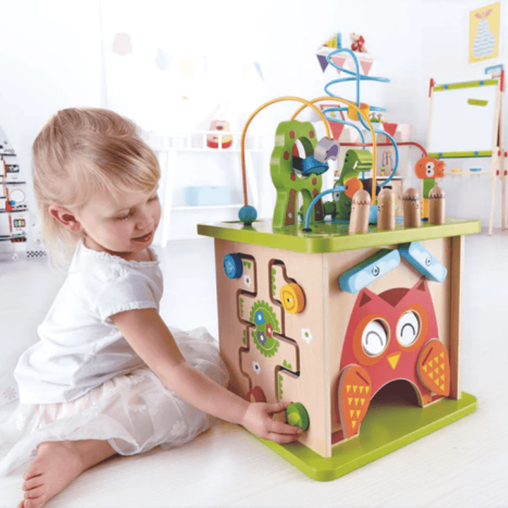 In a playroom, a child engages with the Hape Wildlife Safari Adventure Centre - LUCKY LAST. This colorful educational activity cube is crafted from durable wood and features an engaging owl design, gears, and a bead maze. The toy stimulates cognitive development while turning playtime into an exciting safari adventure.