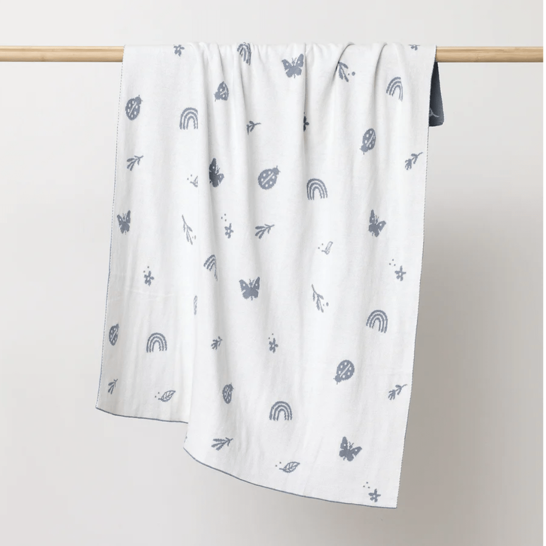 The "Over the Dandelions Organic Cotton Print Blanket" in Enchanted Garden/Sky, featuring delicate gray doodles of rainbows, butterflies, ladybugs, and leaves on a white background, is draped over a wooden rod. It makes an ideal nursery decor piece.