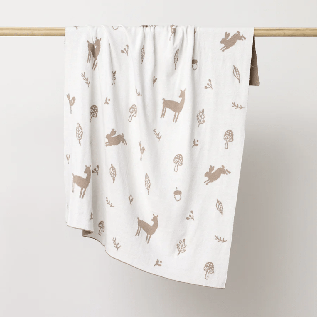 The Over the Dandelions Organic Cotton Print Blanket - LUCKY LAST - ENCHANTED GARDEN/SKY is an exquisite heirloom piece crafted from organic cotton, showcasing delightful brown deer, rabbits, leaves, mushrooms, and acorns on a pristine white background. It elegantly adorns nursery decor, draping beautifully over a wooden rod.