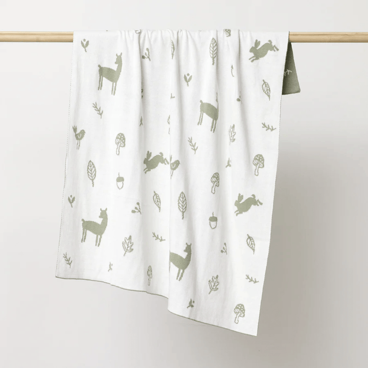 The Over the Dandelions Organic Cotton Print Blanket - LUCKY LAST - ENCHANTED GARDEN/SKY, crafted from organic cotton and adorned with charming patterns of llamas, leaves, and mushrooms in green shades, hangs gracefully on a wooden rod as an enchanting nursery decor piece.