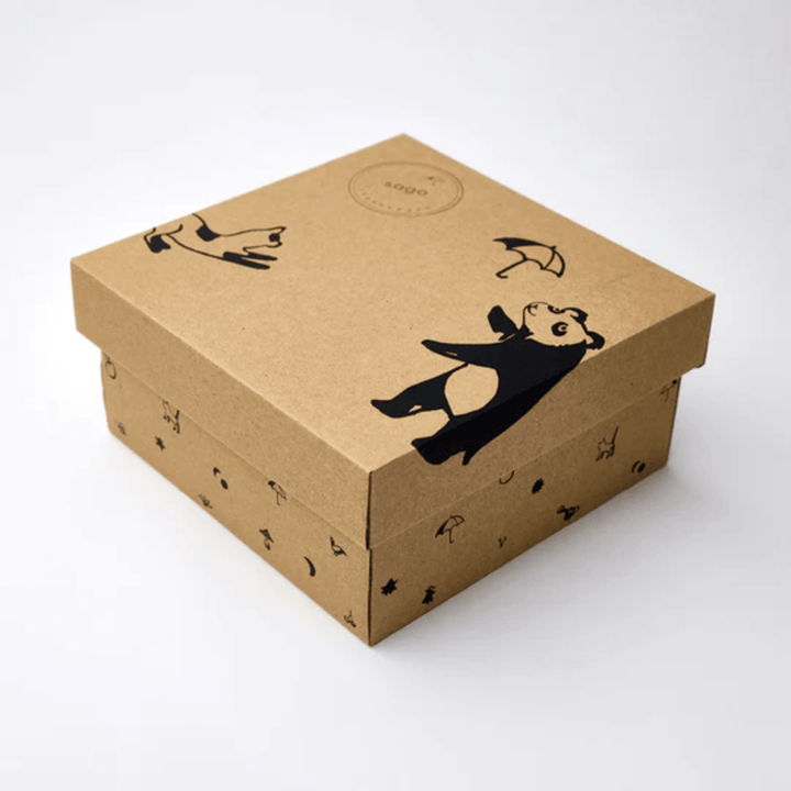 The Saga Copenhagen Gift Box - Elina features a brown cardboard box adorned with cartoon panda and umbrella illustrations on the lid, capturing the charming essence of Saga Copenhagen's gift boxes. This eco-friendly packaging not only brings joy but also aligns with sustainability values through its use of materials that meet organic cotton standards.