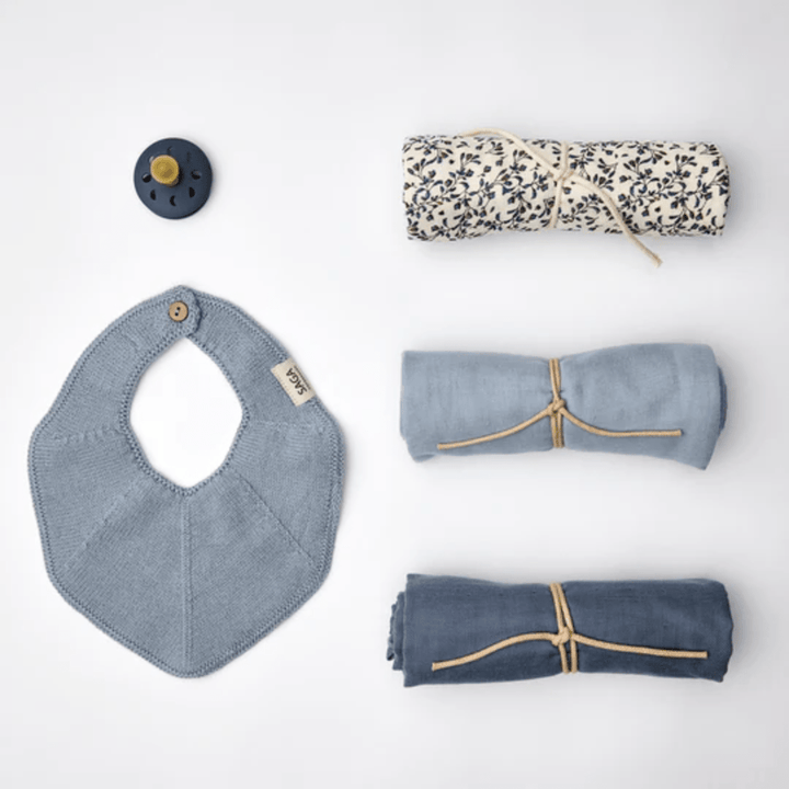 A flat lay of baby items including a pacifier, a cloth bib, and three rolled-up muslin blankets in blue and patterned designs against a white background, perfect for creating an eco-friendly baby gift with luxurious organic cotton baby products from the Saga Copenhagen Snuggle-Up Gift Box by Saga Copenhagen.