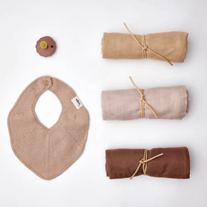 The Saga Copenhagen Snuggle-Up Gift Box, featuring a flower-shaped baby pacifier, a brown knitted baby bib with a button, and four rolled blankets in shades of brown, beige, light beige, and light brown tied with strings, is laid out on a white background—an ideal assortment of organic cotton baby products from Saga Copenhagen.