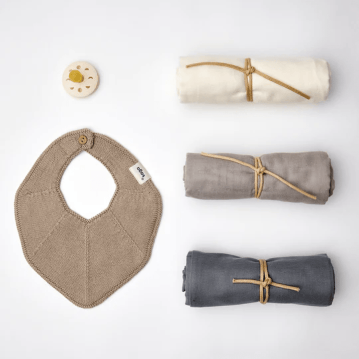 A beige knitted bib, three rolled fabric swaddles tied with string, and a beige pacifier are elegantly arranged on a white background. The Saga Copenhagen Snuggle-Up Gift Box by Saga Copenhagen showcases eco-friendly baby gifts crafted from organic cotton baby products.