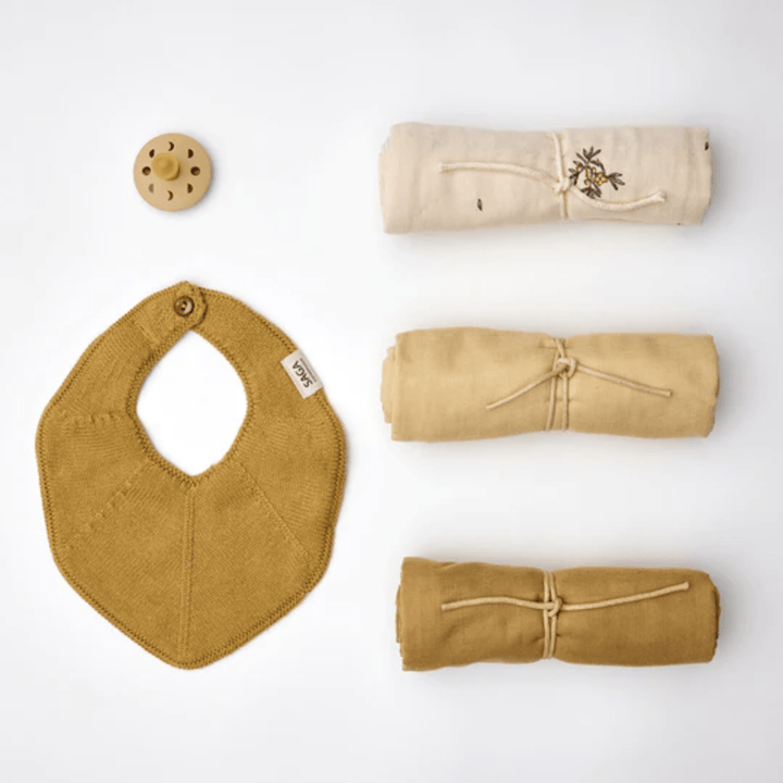 Introducing the delightful Saga Copenhagen Snuggle-Up Gift Box from Saga Copenhagen, which includes a charming yellow bib, a pacifier, and three rolled-up muslin blankets in lovely shades of beige and yellow. Each item is crafted from organic cotton baby products and thoughtfully presented on a pristine white surface.