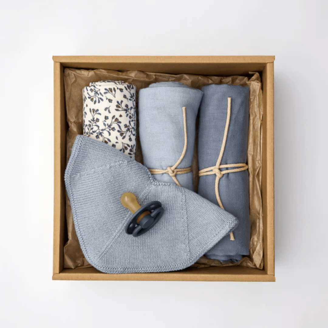 The Saga Copenhagen Snuggle-Up Gift Box from Saga Copenhagen includes a floral-patterned cloth, two rolled blue cloths tied with twine, a folded gray cloth, and a pacifier. This eco-friendly baby gift features organic cotton baby products designed for both comfort and style.