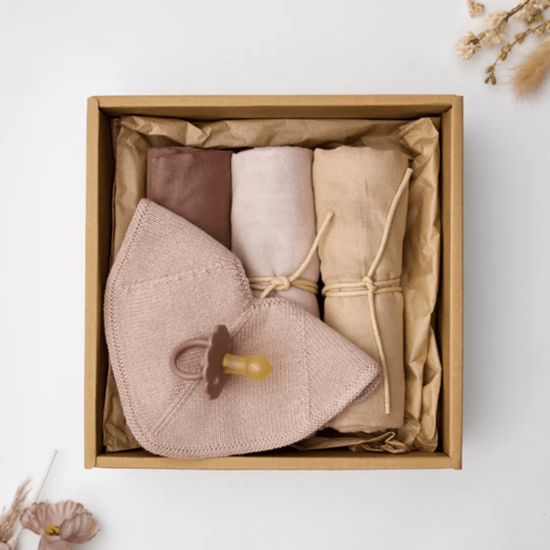 The Saga Copenhagen Snuggle-Up Gift Box by Saga Copenhagen contains three rolled cloths in beige, pink, and brown, along with a pink knit bunny pacifier holder. Made from eco-friendly organic cotton baby products, the box is elegantly surrounded by dried floral elements.