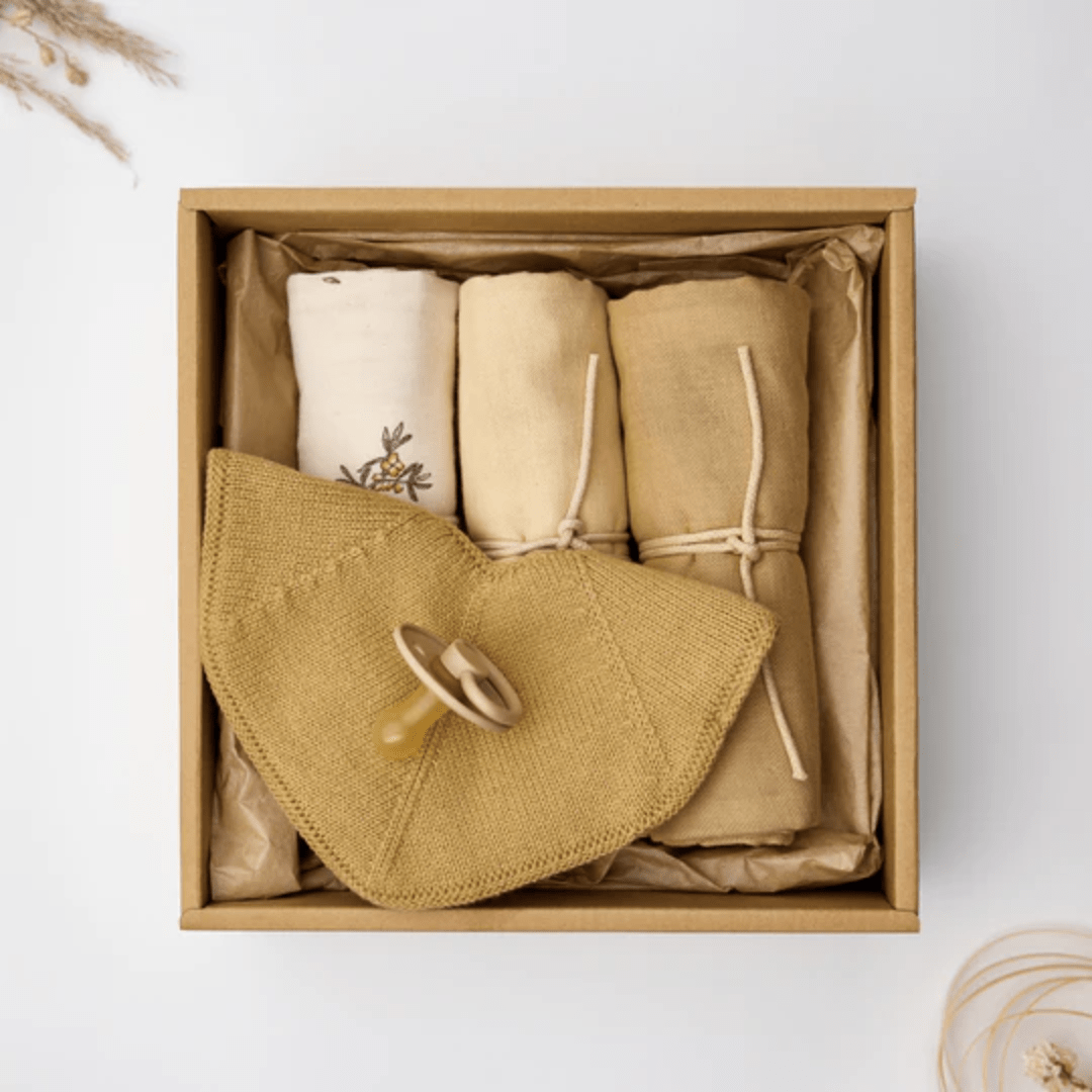 Introducing the Saga Copenhagen Snuggle-Up Gift Box: a luxurious, eco-friendly baby gift featuring an organic cotton pacifier attached to a knitted cloth and three rolled-up blankets in earthy tones, all elegantly arranged on a white background. Perfect for discerning parents who appreciate sustainability.