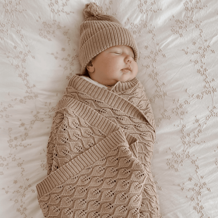 A baby is peacefully sleeping wrapped in an Aster & Oak Organic Leaf Knit Baby Blanket - LUCKY LAST with intricate leaf detailing and wearing a matching brown knitted hat with a pom-pom, lying on a white patterned blanket.