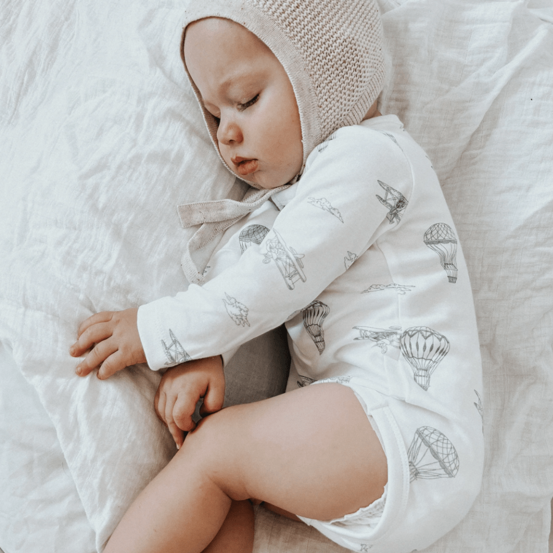 The baby is peacefully asleep, clad in the Aster & Oak Organic Henley Long-Sleeved Onesie from the Fox Woods collection, and a cozy knit bonnet, resting gently on a white pillow.