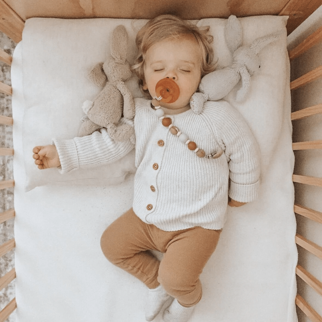 A baby with blond hair sleeps in a crib, wearing a white sweater and brown pants. The baby has a pacifier attached to the Funny Bunny Kids Silicone & Beechwood Dummy Clip and is surrounded by plush bunny toys.