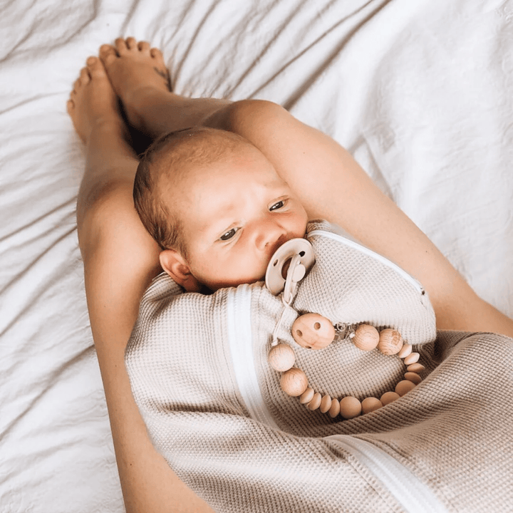 A baby with the Funny Bunny Kids Silicone & Beechwood Dummy Clip, wrapped in a swaddle, rests on a person's legs. The person is lying on a white bedspread.