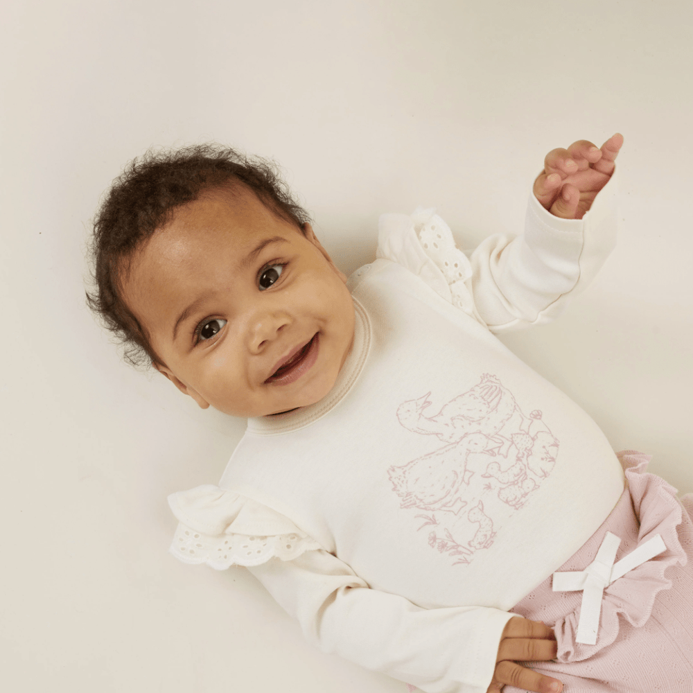 A baby wearing a white long-sleeve top with a pink animal print and frilled sleeves, lying down and smiling. The baby also wears Aster & Oak Organic Pointelle Rib Pale Mauve Leggings made of soft pointelle rib fabric with a cute white bow. Perfect for adding charm to any baby's clothing collection.