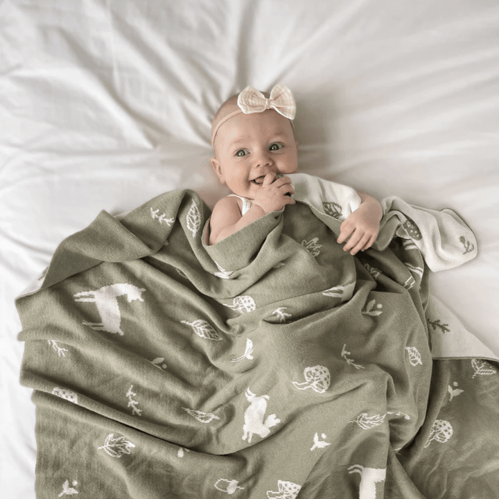 A baby with a bow headband lies on a bed, partially covered by the Over the Dandelions Organic Cotton Print Blanket in the Enchanted Garden/Sky design.