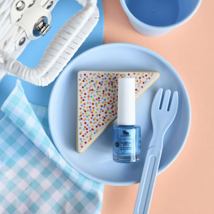 A blue plate with a decorated cookie, a bottle of No Nasties 20 FREE Kids Scented Nail Polish, and a fork and cup on a peach and blue background. A white woven bag and a blue plaid cloth are also visible.