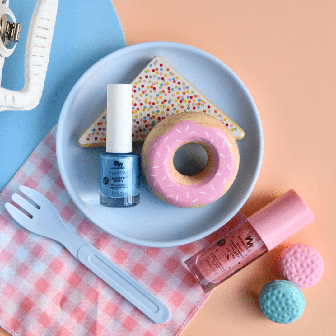 On a pink gingham cloth, a pastel blue plate holds a donut with pink icing, a triangular cookie with sprinkles, and No Nasties 20 FREE Kids Scented Nail Polish in blue and pink. Nearby are a spork, two macaron-shaped items, and the handle of a basket.