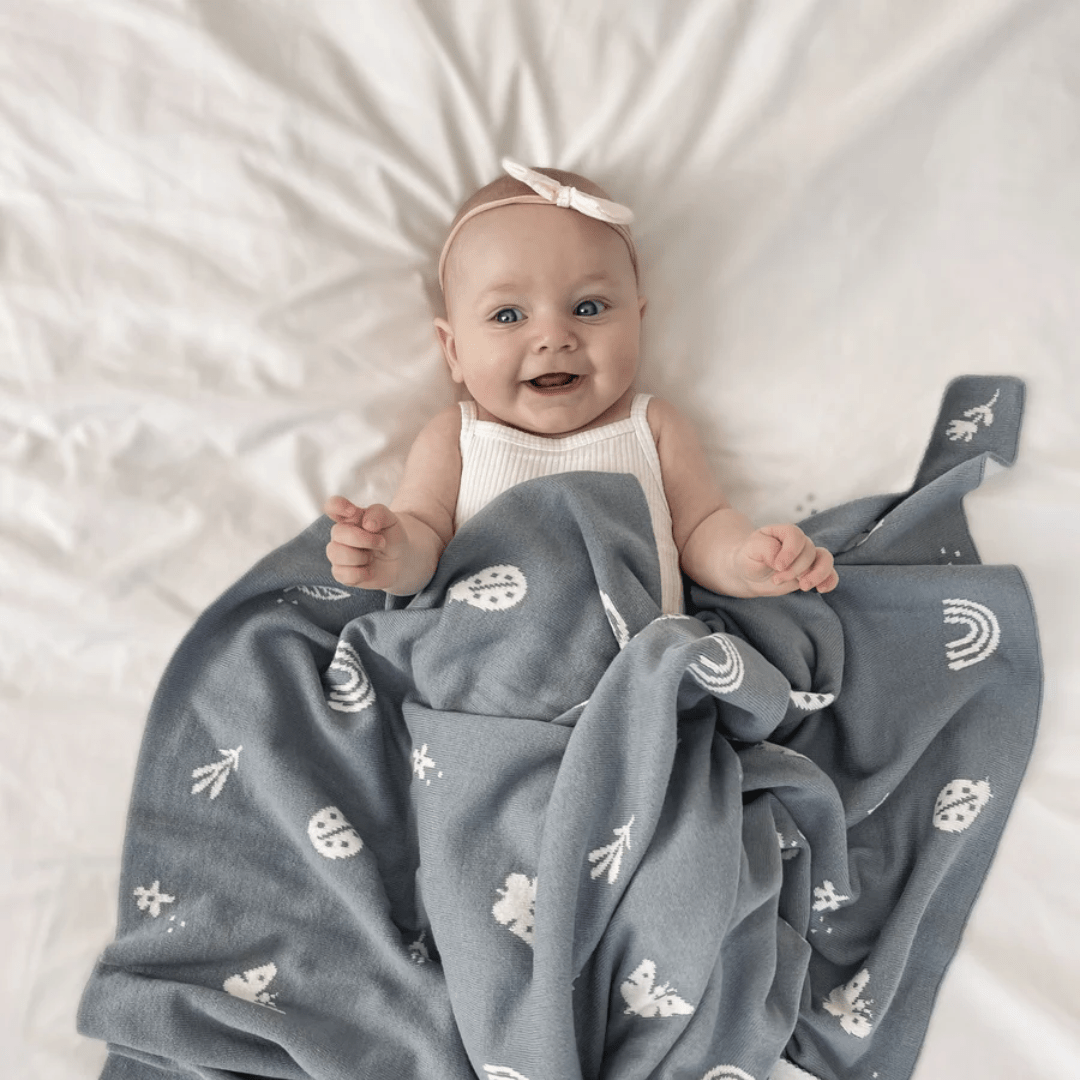 A baby wearing a pink headband and white tank top lies on a white bed with the Over the Dandelions Organic Cotton Print Blanket in Enchanted Garden/Sky, adding a charming touch to any nursery decor.