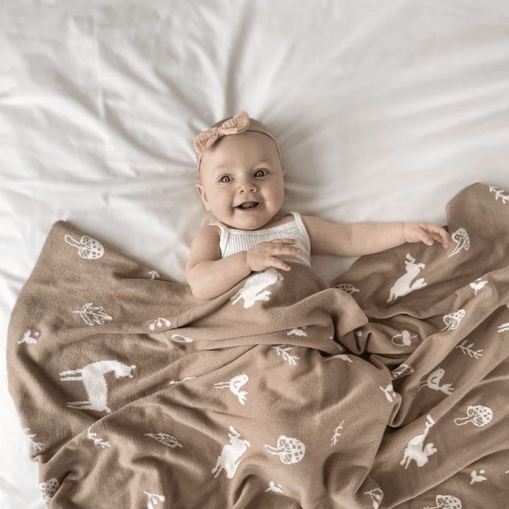 A baby rests on a bed, cozy in the Over the Dandelions Organic Cotton Print Blanket - LUCKY LAST - ENCHANTED GARDEN/SKY, while wearing a pink bow headband.