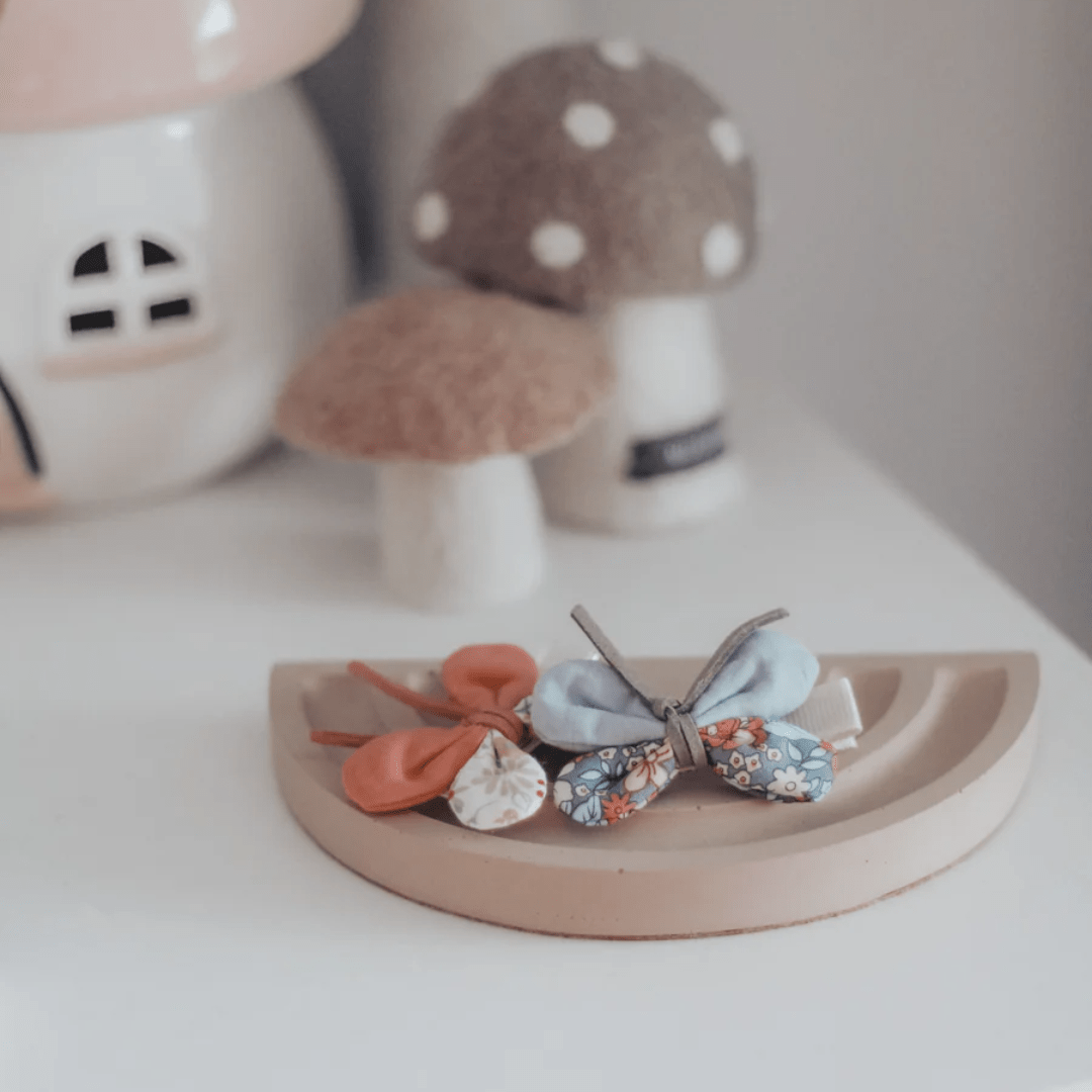 Three Over the Dandelions Butterfly Hair Clips in different colors and patterns are placed on a curved wooden stand, accompanied by decorative mushroom ornaments in the background. This charming setup highlights delightful options for kids' hair accessories.