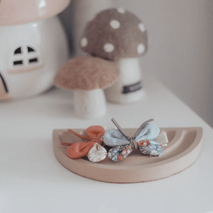 A small wooden tray displays two beautiful Over the Dandelions Butterfly Hair Clips - LUCKY LAST, with one featuring a blue floral design and the other an orange hue. Ideal for kids' hair accessories or toddler hair clips, they create a whimsical vibe alongside the mushroom-shaped ornaments in the background.