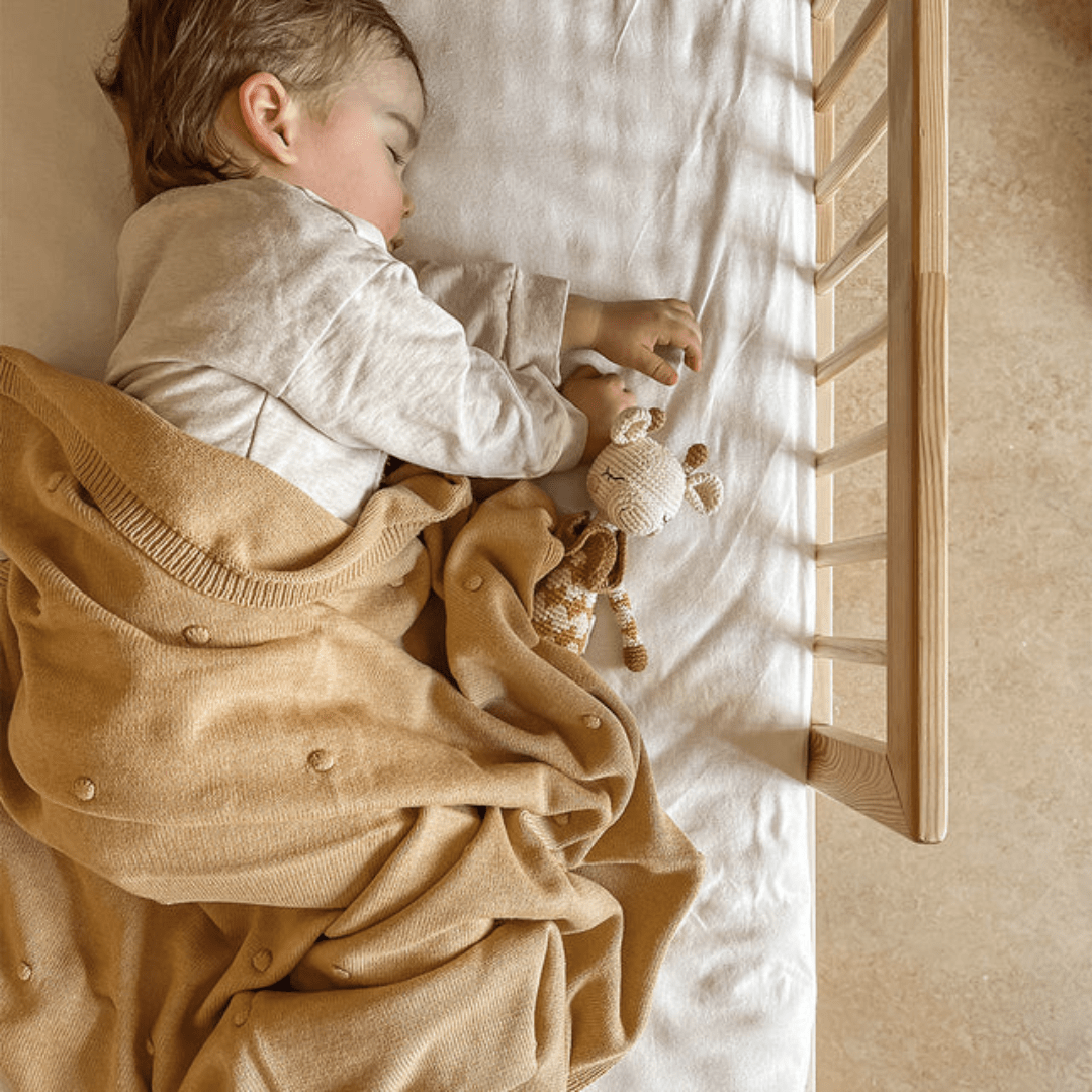 A child sleeps peacefully in a crib, wrapped in a brown blanket and cuddling an eco-friendly toy, the Patti Oslo Organic Cotton Goldie Giraffe.