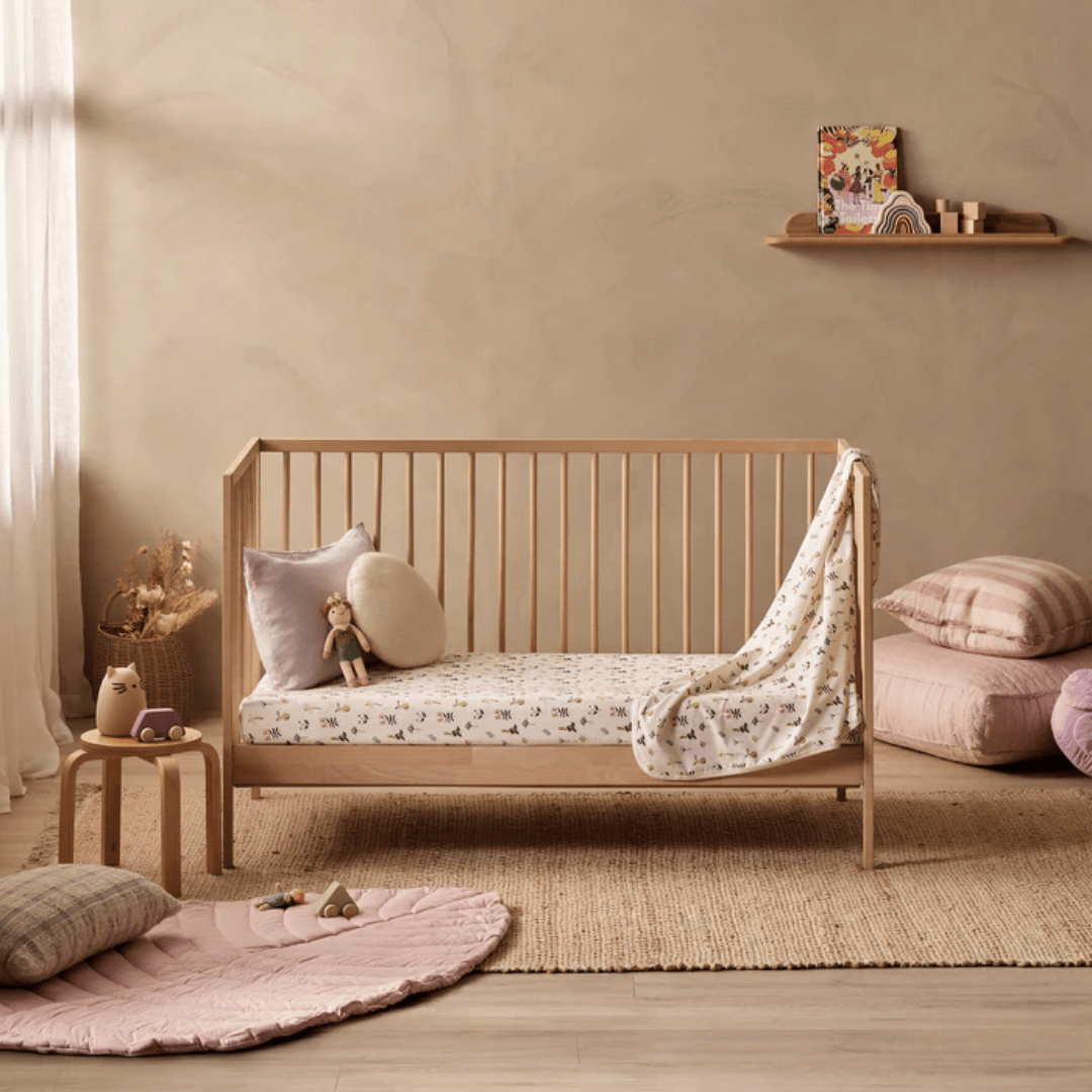 A cozy nursery features a wooden crib with patterned bedding, including the Wilson & Frenchy Organic Cotton Cot Sheet in the exclusive Tribal Woods design, which is GOTS-certified. A small stuffed animal and pillows add charm. A shelf with decor items hangs on the wall, while plush seating and toys are arranged on the floor—an ideal setting for any baby shower gift.