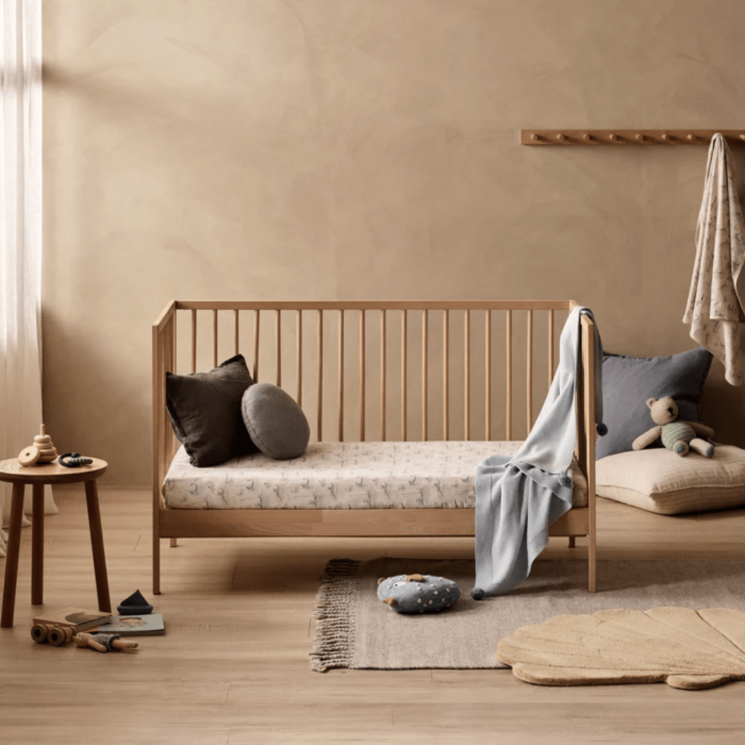 A minimalist nursery showcases a wooden crib adorned with a Wilson & Frenchy Organic Cotton Cot Sheet - Tribal Woods, a small side table holding toys, floor cushions, a rug, and a hook rack with hanging fabric. Neutral tones dominate the decor, making it an ideal setting for a baby shower gift.