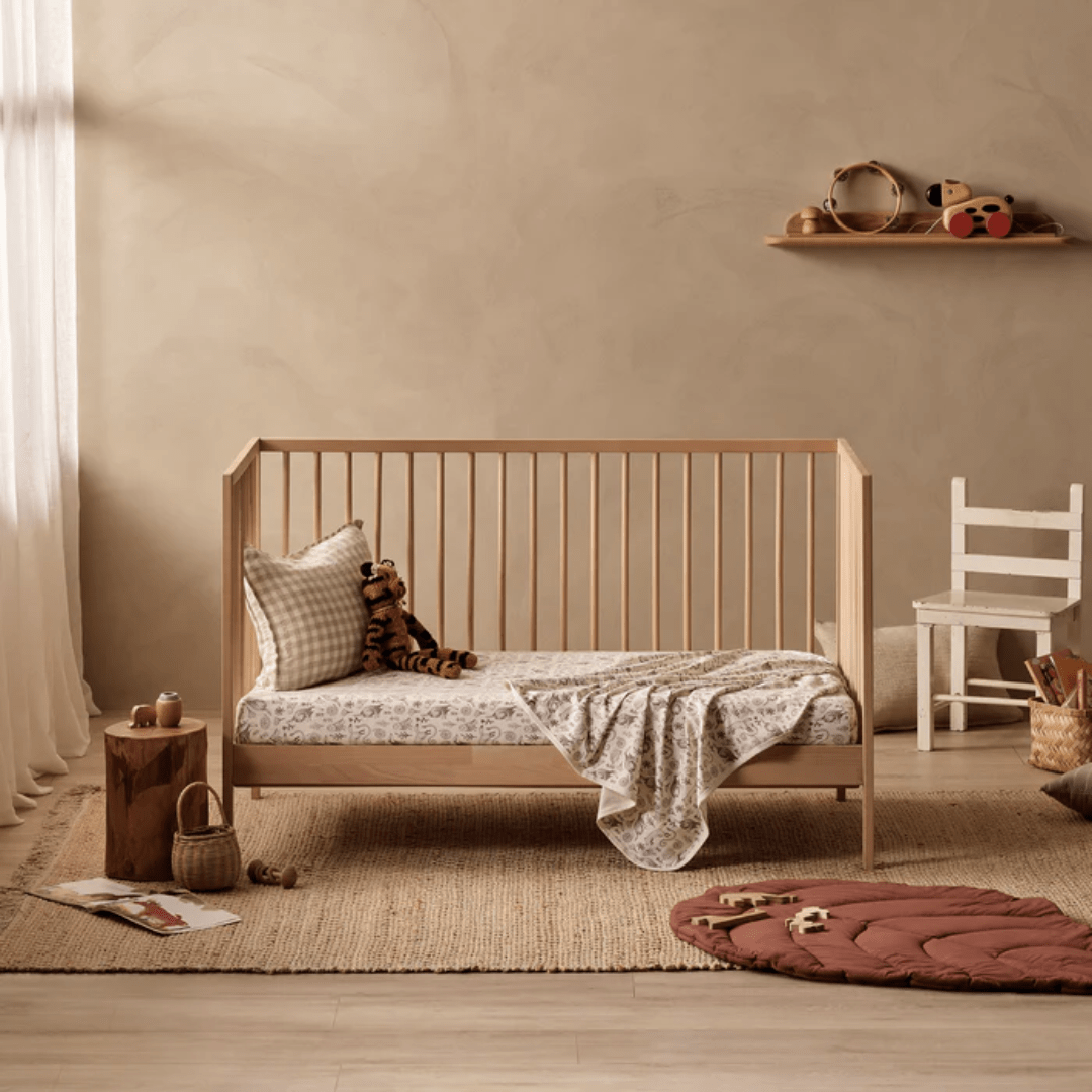 A minimalist nursery features a wooden crib with patterned bedding, the Wilson & Frenchy Organic Cotton Cot Sheet - LUCKY LAST - TRIBAL WOODS ONLY, a plaid pillow, and a stuffed tiger toy. A chair, toys, and decor items are arranged around the room with natural lighting. This setup makes it a perfect Baby Shower Gift idea for new parents.