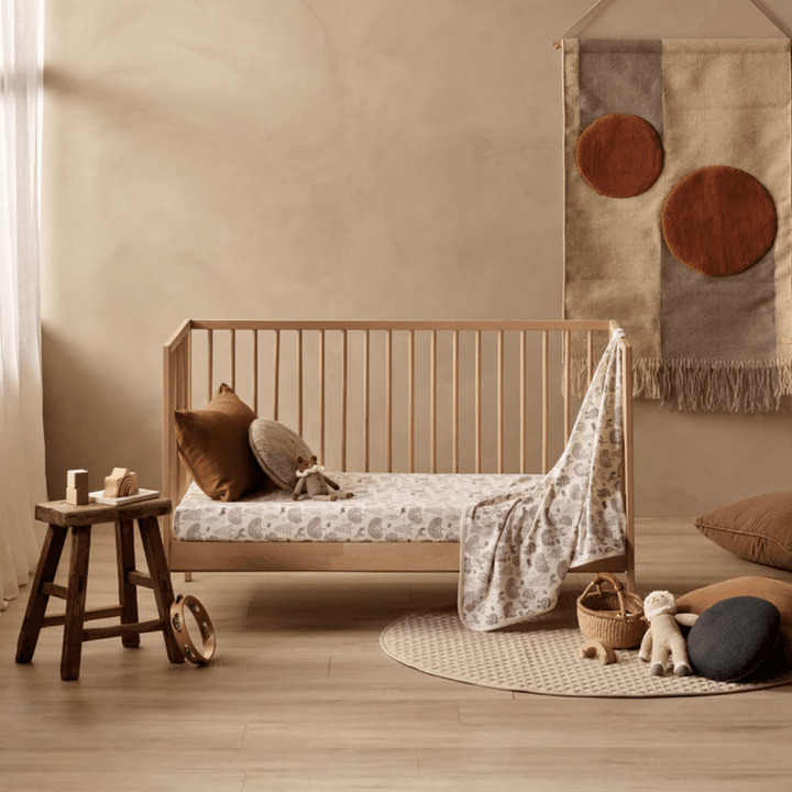 A minimalist nursery featuring a wooden crib dressed with the Wilson & Frenchy Organic Cotton Cot Sheet in the exclusive Tribal Woods pattern. Neutral bedding complements a small stool, cushions, toys, and a beige wall hanging adorned with red and brown circles. Natural light filters through a sheer curtain, creating an ideal setup that could also serve as the perfect baby shower gift.