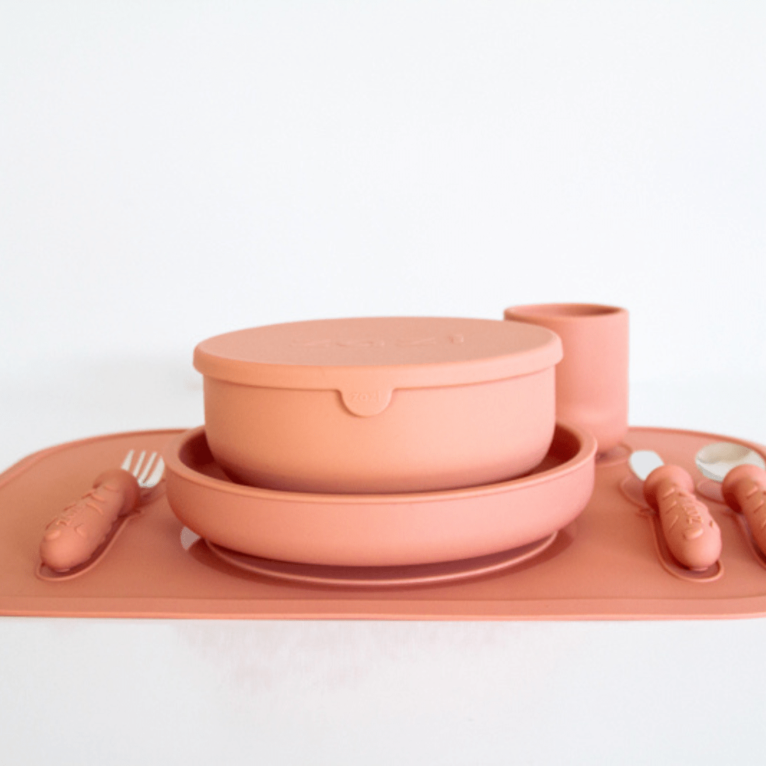 A Zazi Clever Mat children's dining set in mauve silicone, including a bowl with lid, a plate, a cup, utensils, and the Clever Mat with integrated suction technology by Zazi, all displayed neatly on a white background.