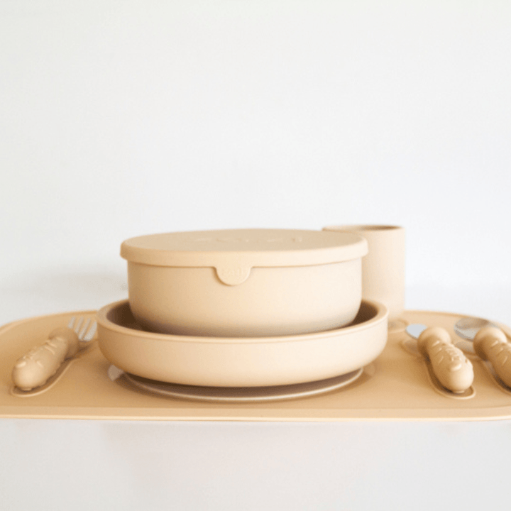 A set of beige-colored plastic dinnerware, including plates, bowls with lids, cups, and utensils, arranged neatly on a coordinating Zazi Clever Mat from the brand Zazi against a plain white background.