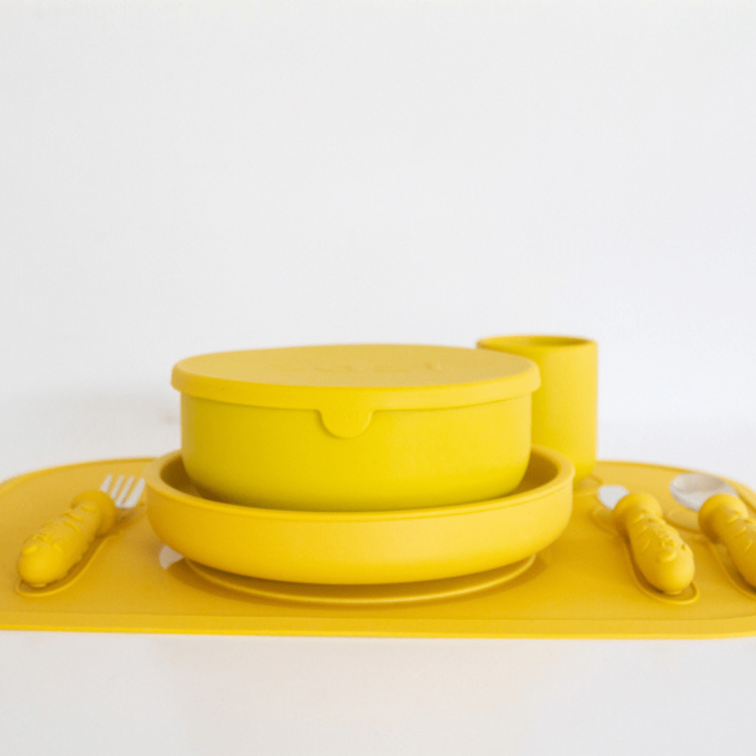 A set of yellow tableware, crafted from food-grade silicone, including a bowl with a lid, a plate, utensils, and a cup arranged on a Zazi Clever Mat by the brand Zazi against a white background.