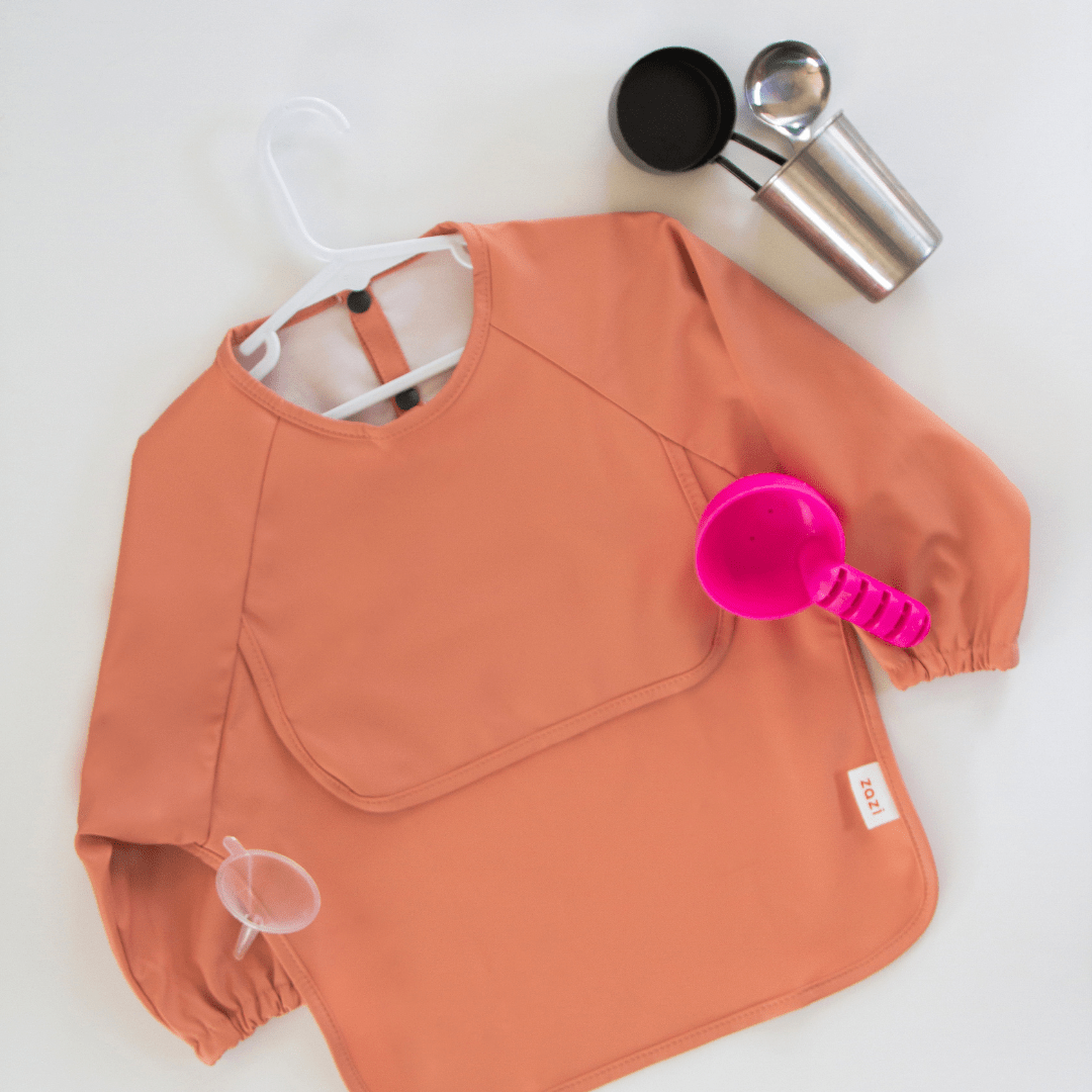 A Zazi Recycled Full-Sleeved Bib in peach, a bright pink scoop, a metal scoop, a metal container, and a white hanger are arranged on a white surface.