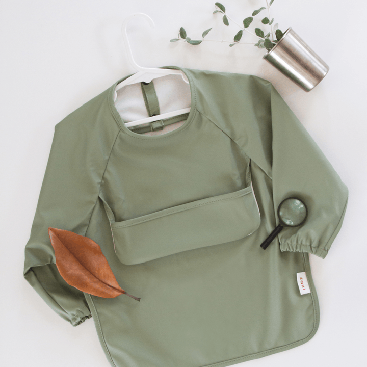 A Zazi Recycled Full-Sleeved Bib in green is laid flat on a white surface, accompanied by a magnifying glass, a brown leaf, and a small metal cup holding a plant.