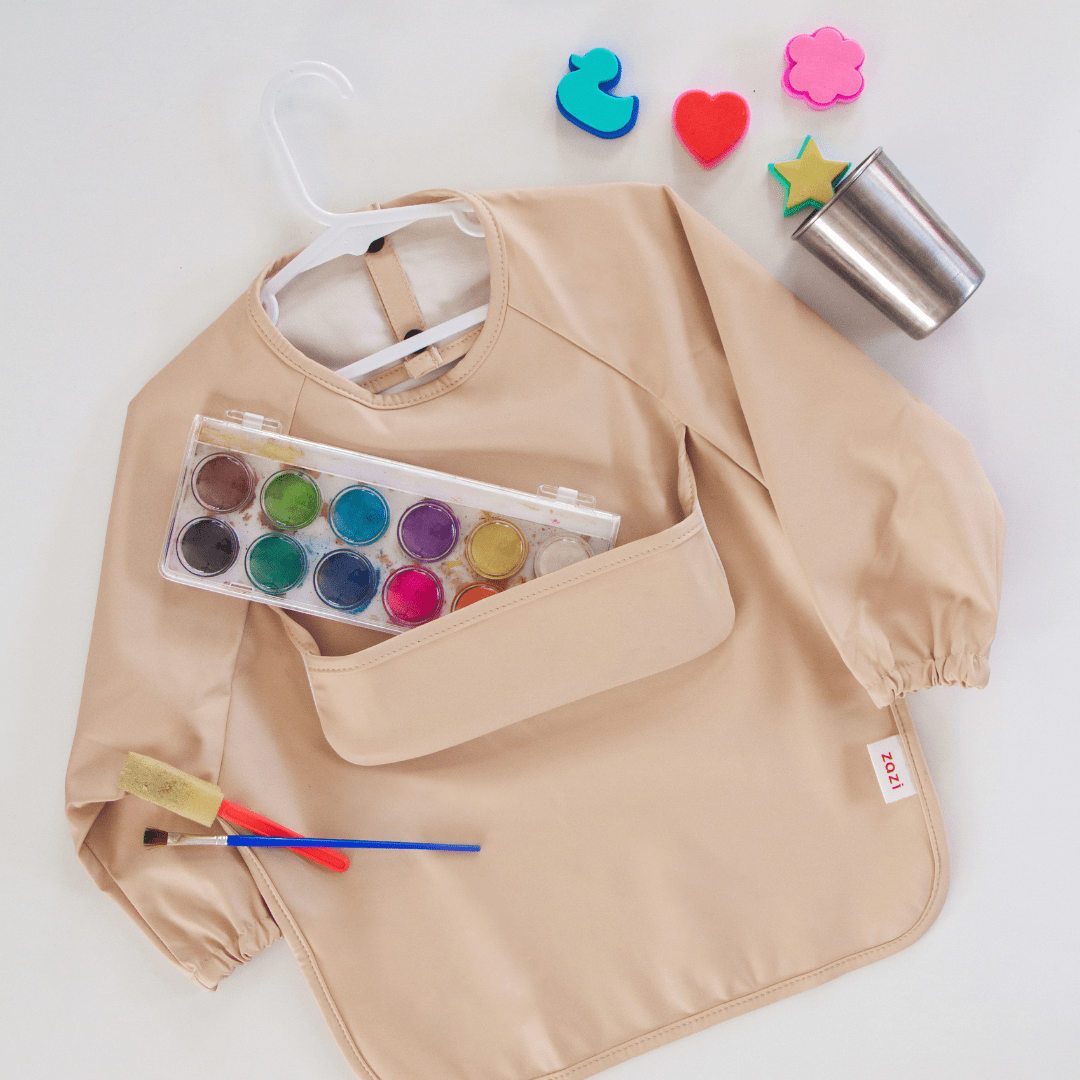 Beige smock with a front pocket, paint palette with twelve colors, paintbrush, sponge brush, metal cup, blue rubber duck, pink flower-shaped cutout, star-shaped sponge, and heart-shaped sponge. Also includes machine washable Zazi Recycled Full-Sleeved Bibs from Zazi to keep little artists clean.