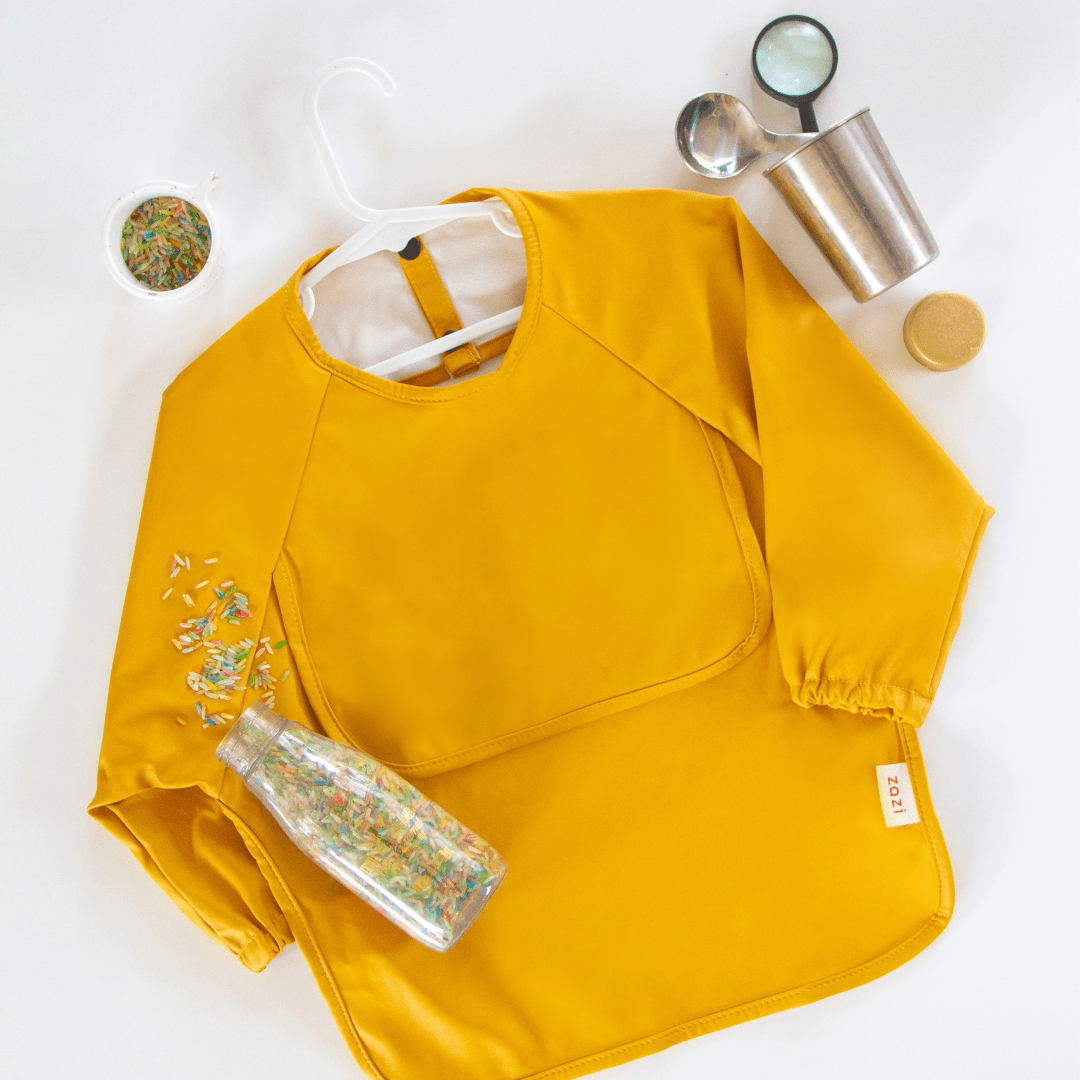 A yellow Zazi Recycled Full-Sleeved Bib, a bottle of rice, a scoop, and a measuring cup are arranged on a white surface.