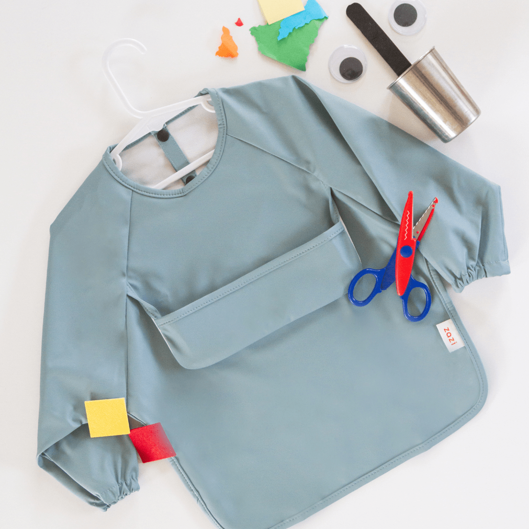 A Zazi Recycled Full-Sleeved Bib in light blue, featuring long sleeves and a front pocket, is laid out on a white surface. Nearby are a pair of scissors, craft paper pieces, googly eyes, and a metal cup with black felt pieces.