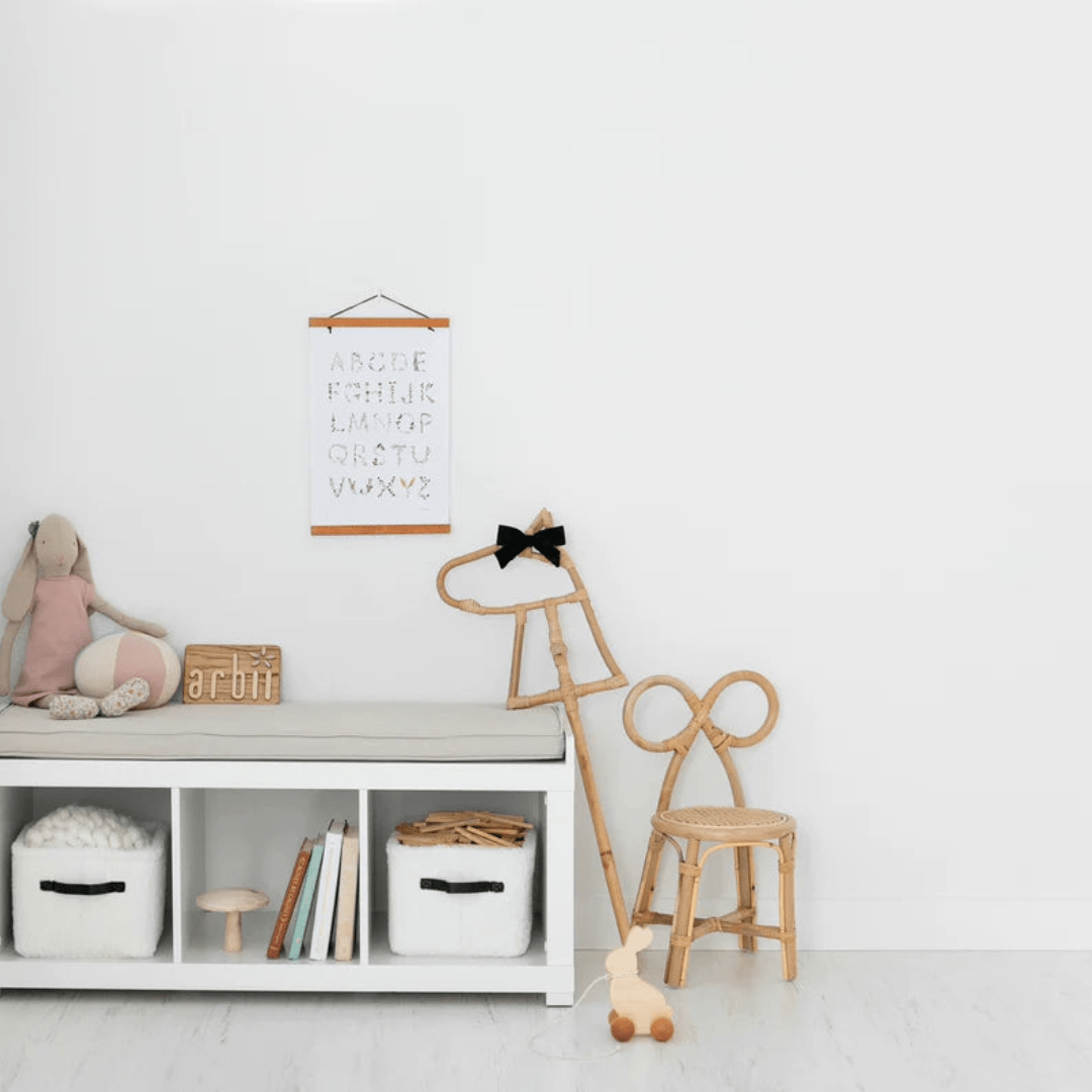 A minimalist children’s room features a white shelf filled with toys and books, a plush rabbit, and a decorative alphabet print on the wall. Wooden toys and furniture foster imaginative play, highlighted by the delightful Classical Child Rattan Hobby Horse - LUCKY LAST, which adds an eco-friendly touch.