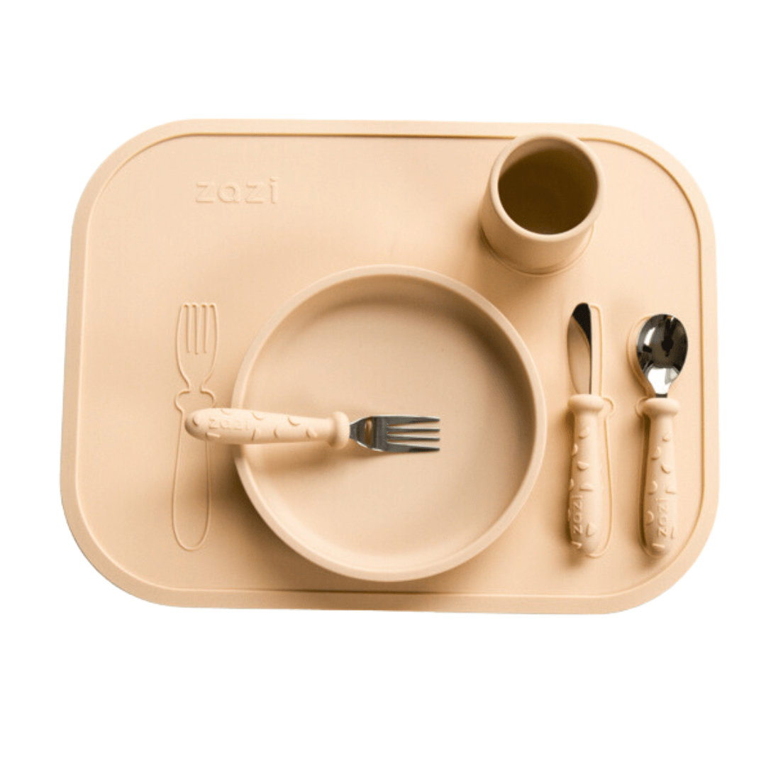 Zazi children's dining set including a plate, cup, fork, spoon, and knife arranged on a Zazi Clever Mat. The items are in beige color with textured, kid-friendly handles made of food-grade silicone.