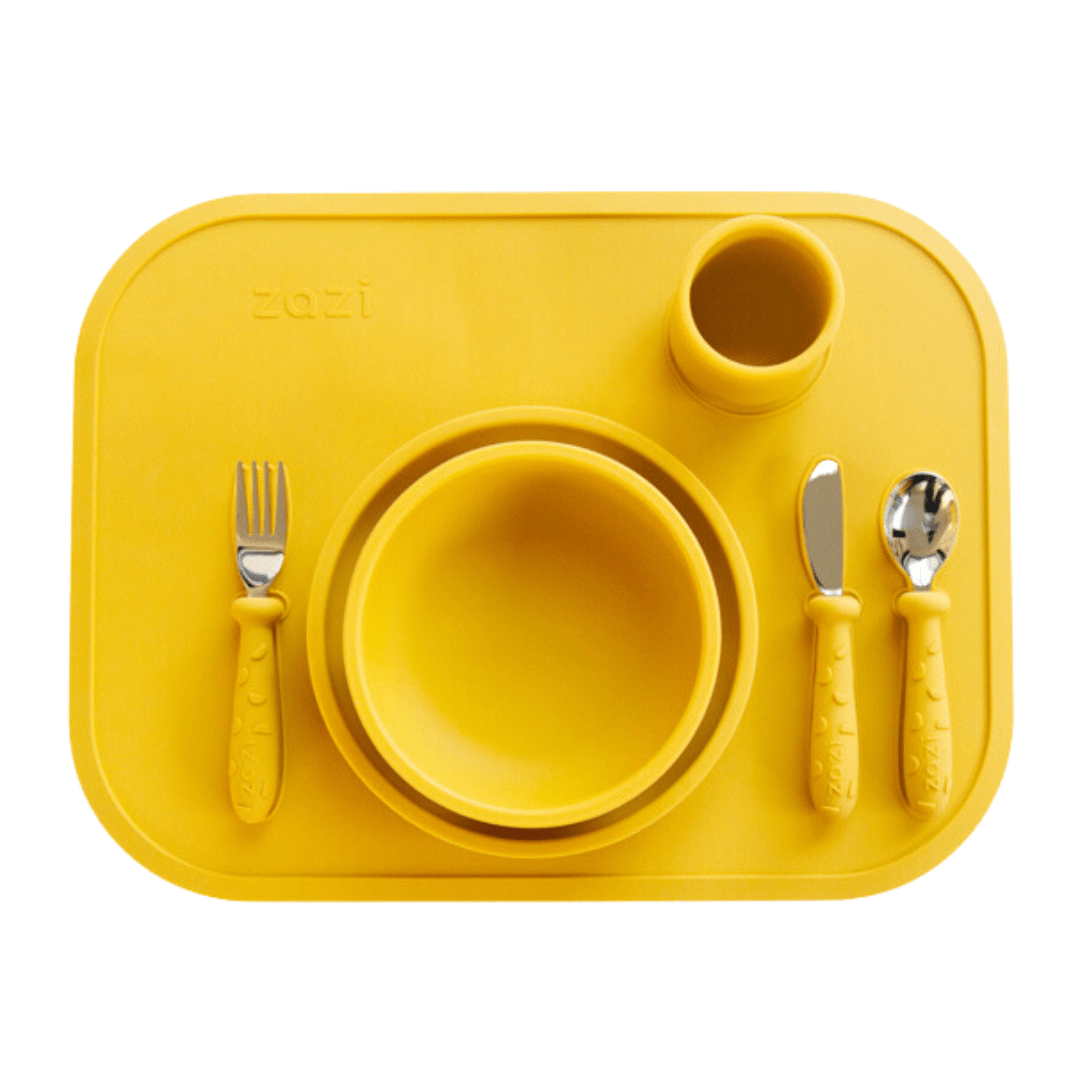 A Zazi Clever Mat in a vibrant yellow, crafted from food-grade silicone, includes a bowl, plate, cup, fork, knife, and spoon. Each item is neatly arranged in designated spaces on the mat with innovative suction technology to keep everything secure.