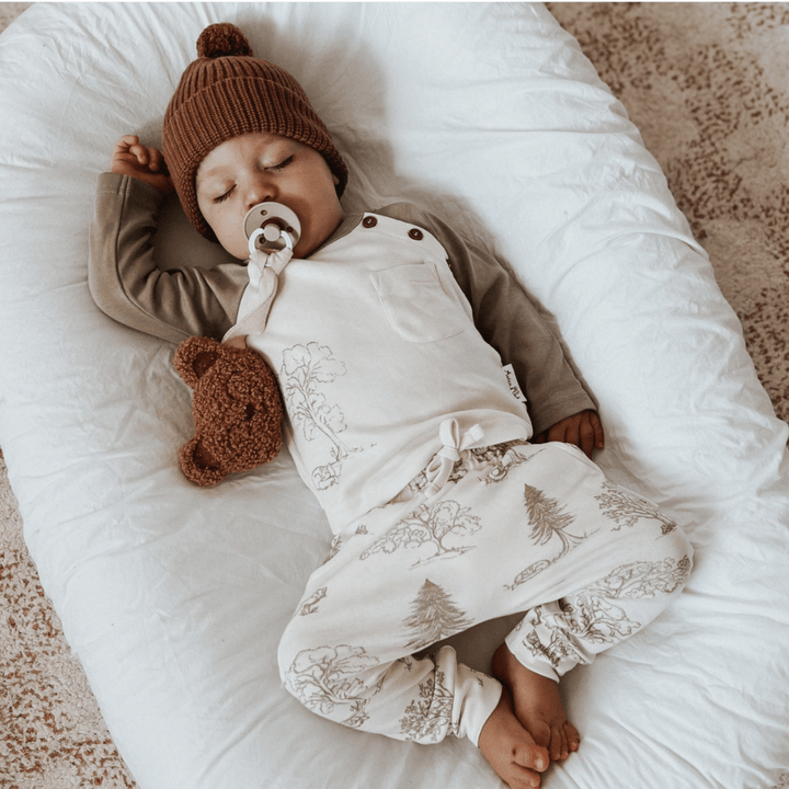 A baby resting on a white cushion is wearing a printed outfit and brown knit hat from Aster & Oak, clinging to a brown teddy bear while using a pacifier. The cozy outfit includes the Aster & Oak Organic Harem Pants - LUCKY LASTS, crafted from GOTS-certified organic cotton, offering comfort and sustainability.