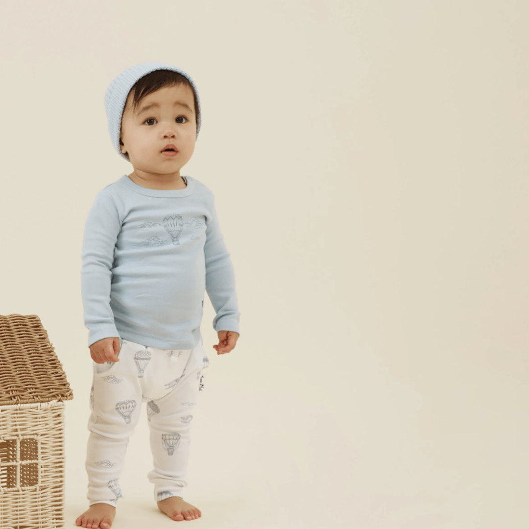 A toddler in a blue hat and long-sleeve shirt, paired with Aster & Oak Organic Harem Pants made from GOTS-certified organic cotton, stands beside a wicker basket on a light background.