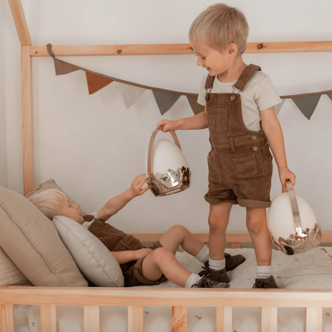 Two boys in overalls playing on a bed with Little Belle Nightlights Dragon Egg Carry Lantern nightlights.